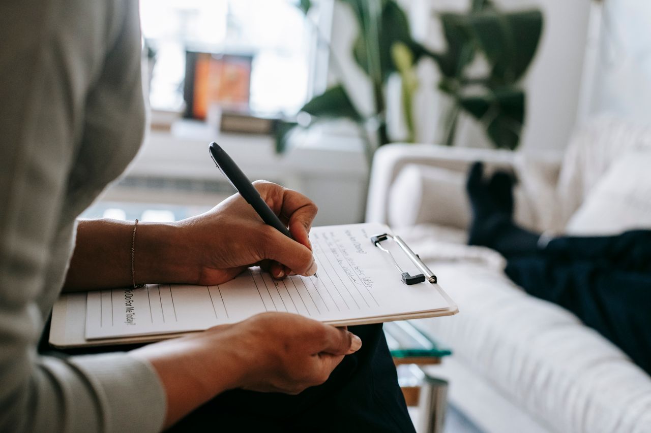 heros-img Unrecognizable ethnic female therapist taking notes on clipboard while filling out form during psychological appointment with anonymous client lying on blurred background